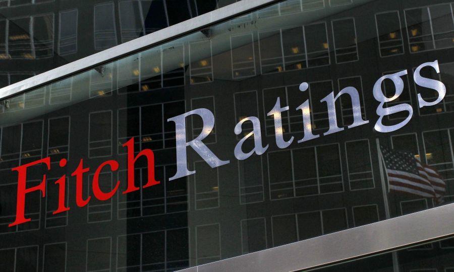 A close-up, angled shot of the "Fitch Ratings" logo displayed on a glass window. The glass reflects a dark office building with many windows and an American flag flying in the background.